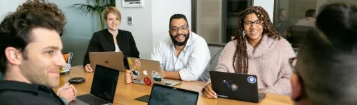 Two women and two men in a meeting listening to someone else speaking, all with laptops around a table